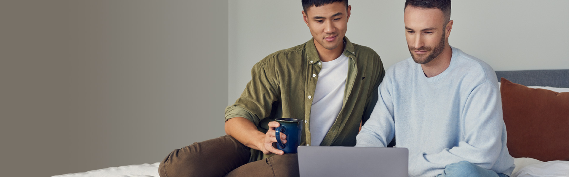 Zwei Männer sitzen entspannt auf einem Bett und schauen auf einen Laptop.